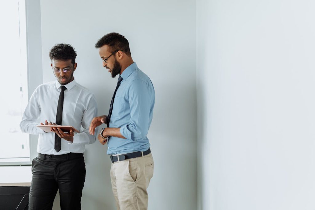 Two Men in Formal Clothes and Eyeglasses Holding Tablet Beside White Wall